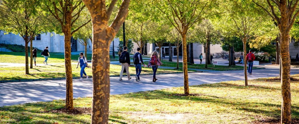 An image of campus with students walking outside on a sunny day on the sidewalks. 