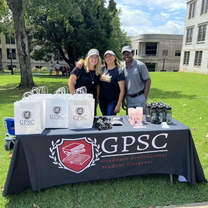 A picture of 3 graduate students at a GPSC table on the lawn of the university in front of Old Main giving out gifts to students.