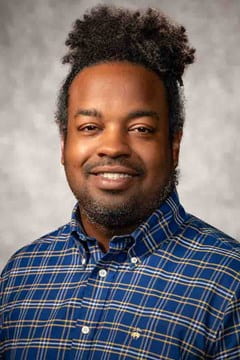 An image of a black male with a plaid shirt and dark long curly hair pulled back.