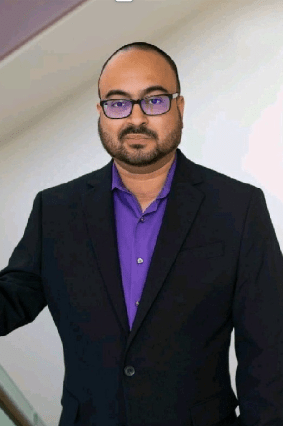 An image of a latino male with glasses in a black blazer and purple shirt. 